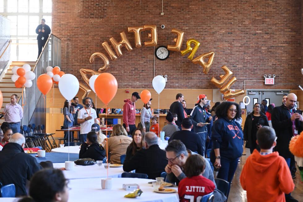一大群人在菲尔德斯顿伦理文化学校的餐厅里坐着站着, 周围是橙色和白色的气球，还有 